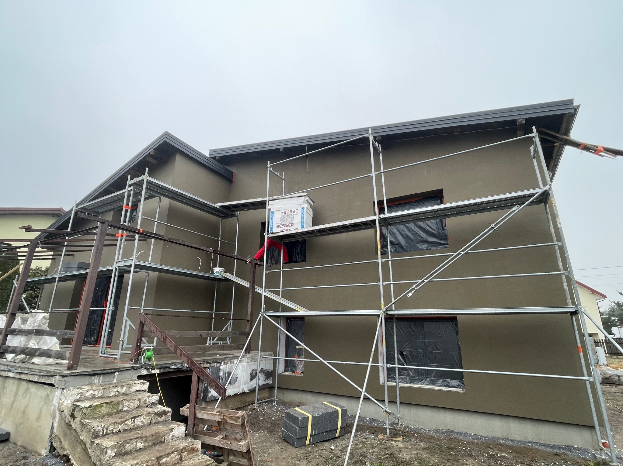 Dom jednorodzinny w trakcie ocieplania, widoczne rusztowania, okna zabezpieczone folią, paczka styropianu na rusztowaniu, elewacja w kolorze beżowym.
