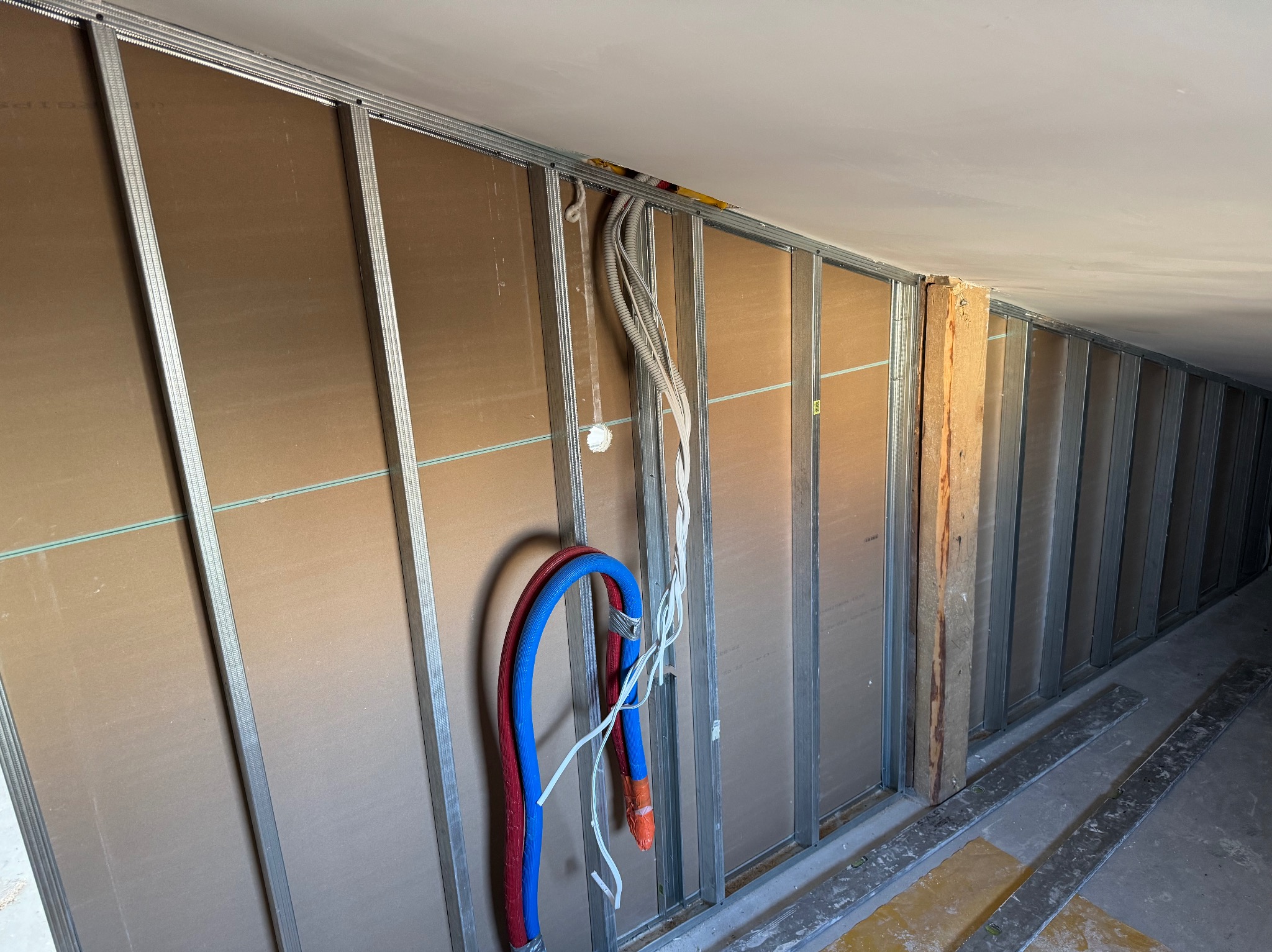 Konstrukcja ścian działowych z profili stalowych i płyt, widoczne przewody instalacji elektrycznej i rury instalacji wodnej, fragment podłogi z żółtymi plamami.