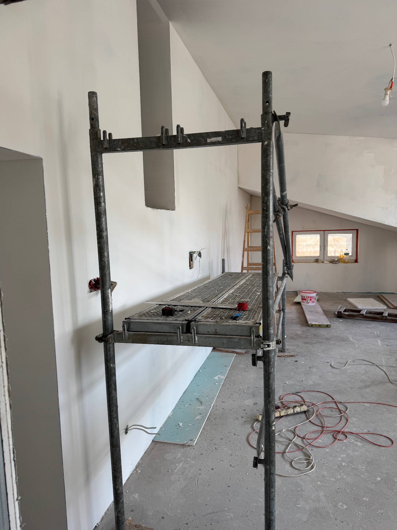 Wnętrze w trakcie prac wykończeniowych: metalowe rusztowanie, ściany po szpachlowaniu, widoczne puszki elektryczne, podłoga z resztkami materiałów budowlanych, okno dachowe w tle.