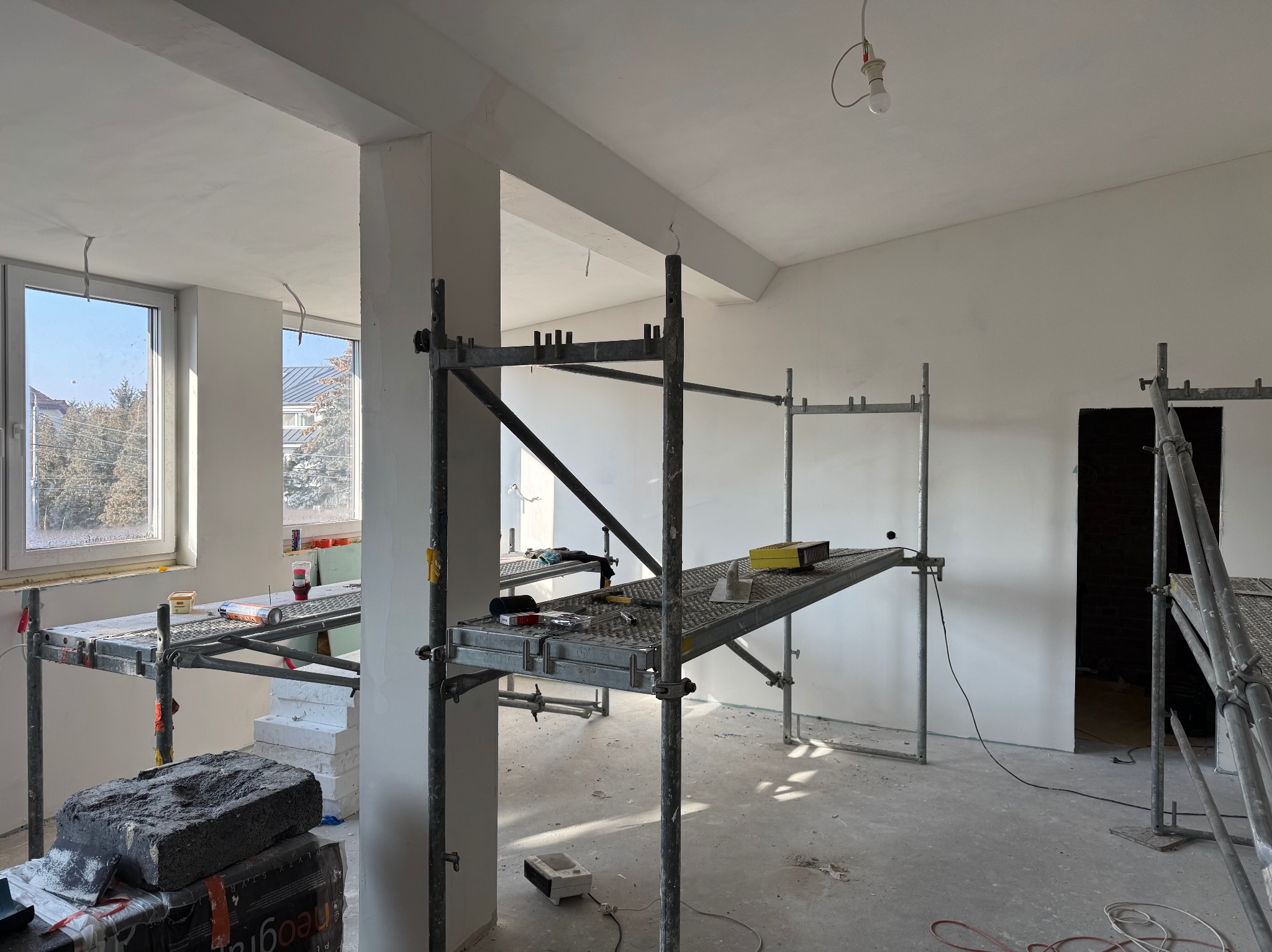 Wnętrze w trakcie prac wykończeniowych: rusztowania, narzędzia, materiały budowlane, ściany przygotowane pod malowanie lub gładzie, widok przez okno na zewnątrz.