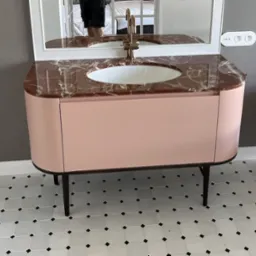 Różowa szafka łazienkowa z marmurowym blatem i złotą armaturą, odbicie osoby w lustrze, biała mozaika podłogowa.
