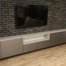 MR-STYL MEBLE NA WYMIAR - Długa, szara szafka RTV podwieszona pod telewizorem na ścianie z cegły dekoracyjnej, widok z perspektywy podłogi.