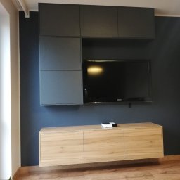 MR-STYL MEBLE NA WYMIAR - Nowoczesna aranżacja ściany telewizyjnej z szarymi szafkami wiszącymi i podwieszaną szafką RTV z frontami w kolorze jasnego drewna.