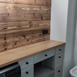 MR-STYL MEBLE NA WYMIAR - Szara szafka łazienkowa z drewnianym blatem i krzesłem w pepitkę, umieszczona obok pralki na tle ściany z desek i lampy.