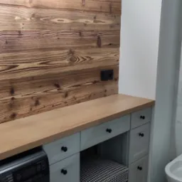 Szara szafka łazienkowa z drewnianym blatem i krzesłem w pepitkę, umieszczona obok pralki na tle ściany z desek i lampy.