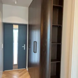 Wysoka, ciemna szafa wnękowa z otwartymi półkami na końcu, z minimalistycznymi, pionowymi uchwytami, w przedpokoju z parkietem w jodełkę i szarymi drzwiami.