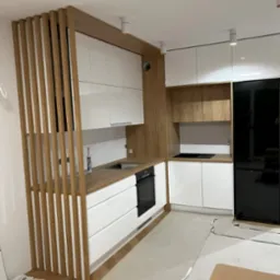 Nowoczesna kuchnia z białymi szafkami i drewnianymi akcentami, widoczna drewniana ścianka działowa z pionowych listew, czarna lod&oacute;wka i niedokończona podłoga.