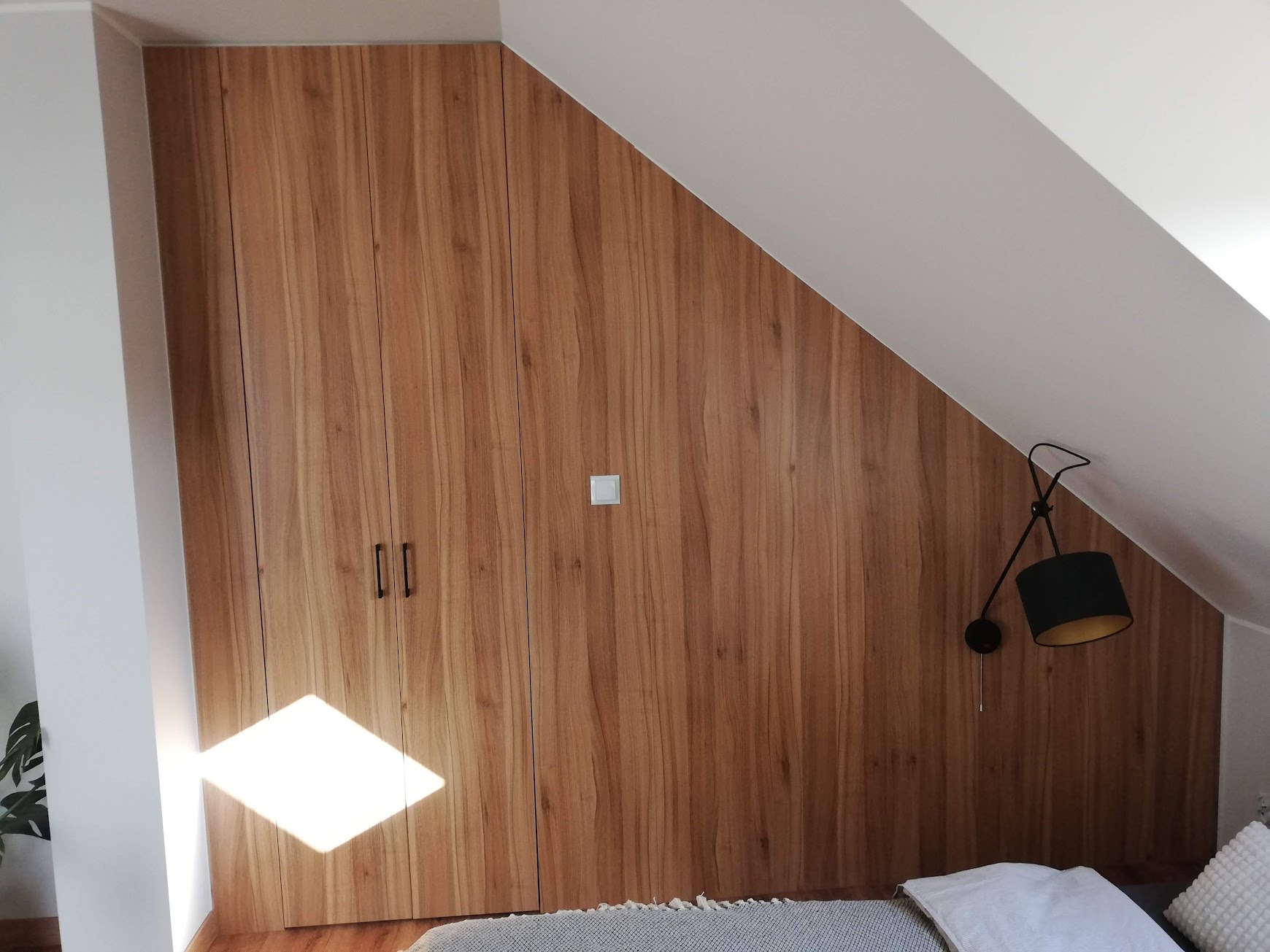 Drewniana szafa wnękowa z oświetleniem ściennym w pokoju na poddaszu, widoczny fragment łóżka z pościelą.