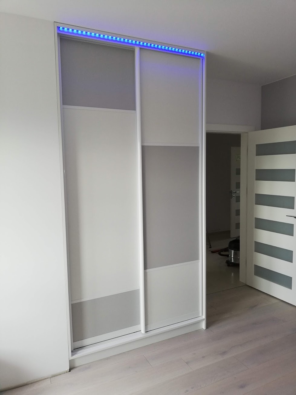 Biała szafa wnękowa z przesuwanymi drzwiami, podświetlona niebieskim paskiem LED, w jasnym pokoju z drewnianą podłogą.