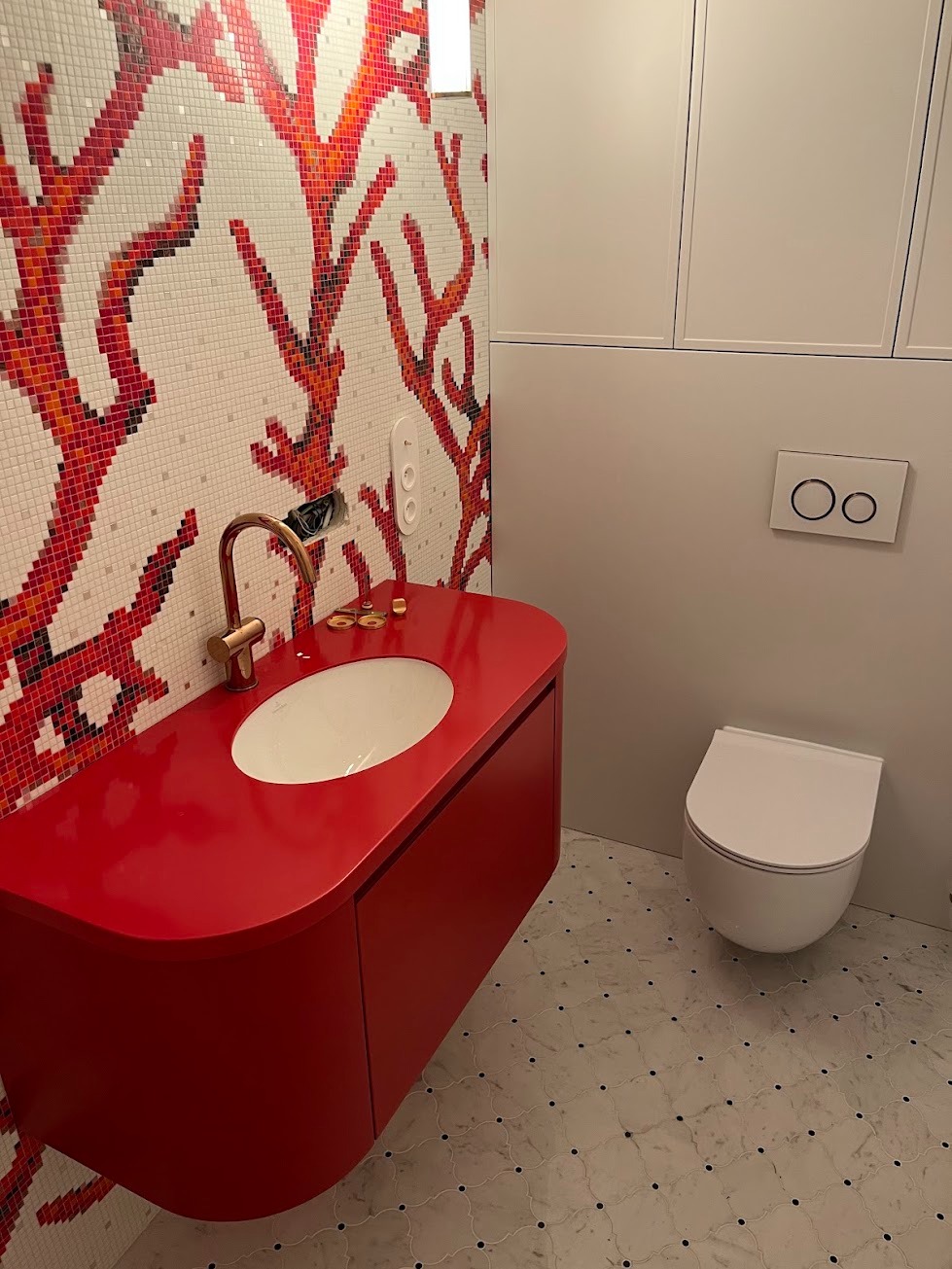 Nowoczesna łazienka z czerwoną szafką pod umywalką, złotą baterią i mozaiką koralową na ścianie, obok wisząca toaleta.