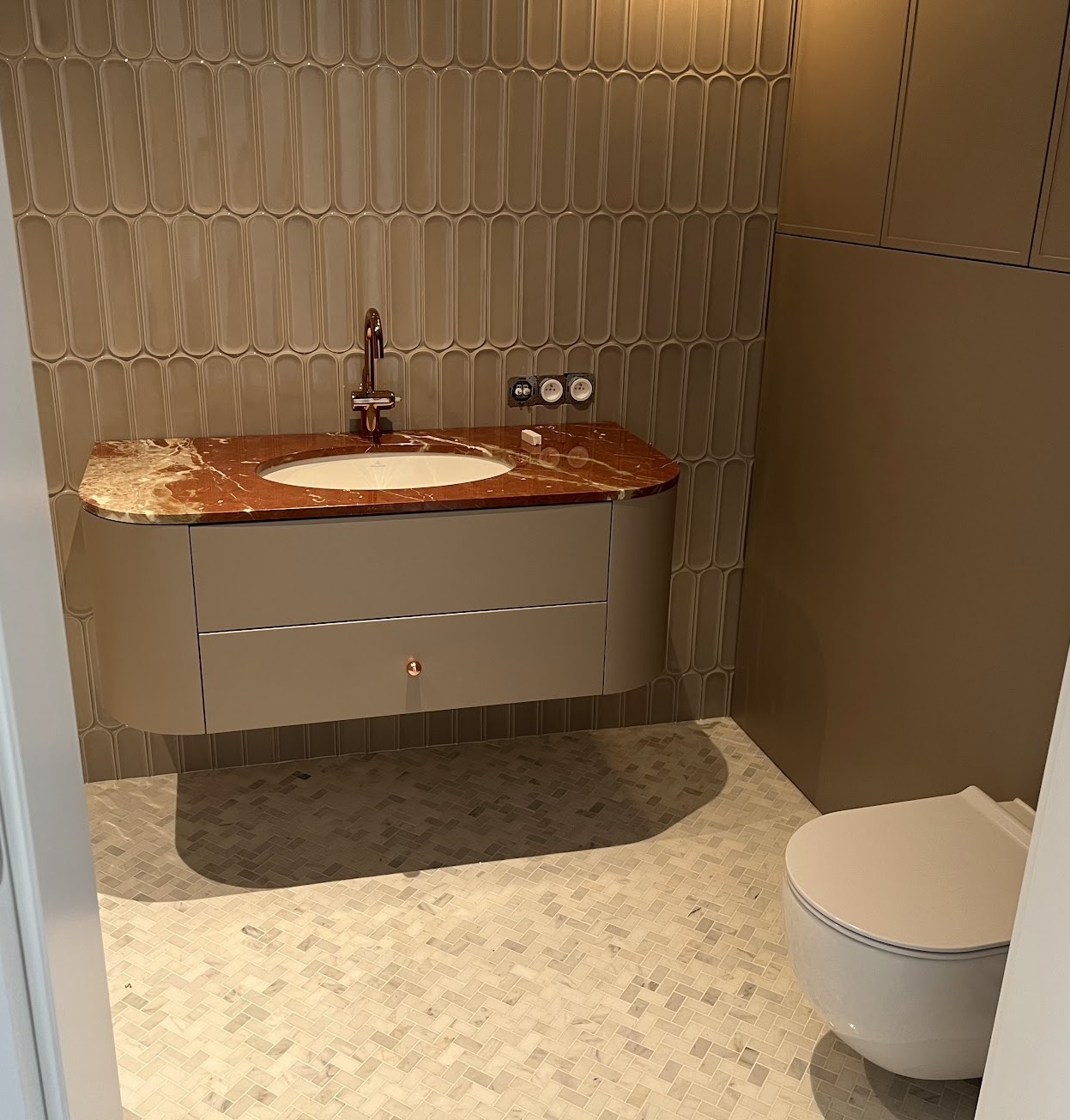 Nowoczesna łazienka w beżach: podwieszana szafka z umywalką i blatem z czerwonego marmuru, owalne płytki na ścianie, mozaika na podłodze, wisząca toaleta.