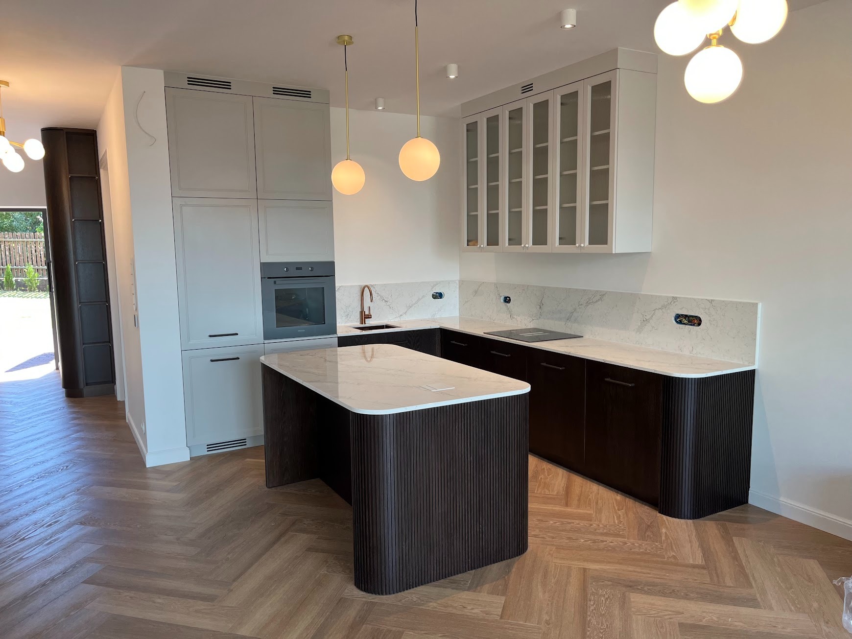 Nowoczesna kuchnia z wyspą, fronty szafek w kolorze ciemnego drewna z ryflowaną strukturą, marmurowy blat i backsplash, podłoga w jodełkę, złote lampy wiszące.