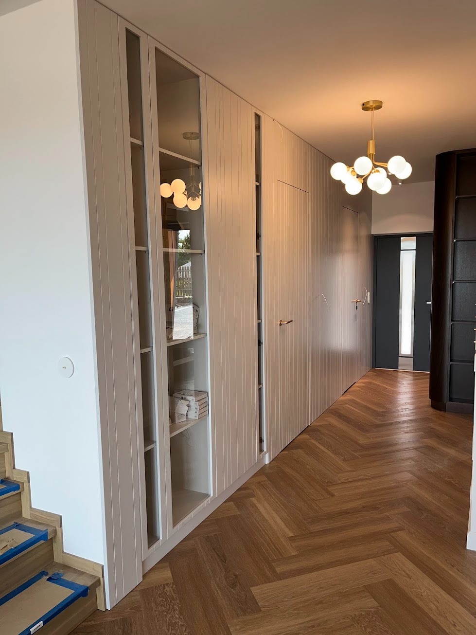 Korytarz z zabudową szaf wnękowych z białymi frontami z pionowymi frezowaniami i przeszklonymi witrynami, parkiet ułożony w jodełkę, nowoczesna lampa sufitowa.