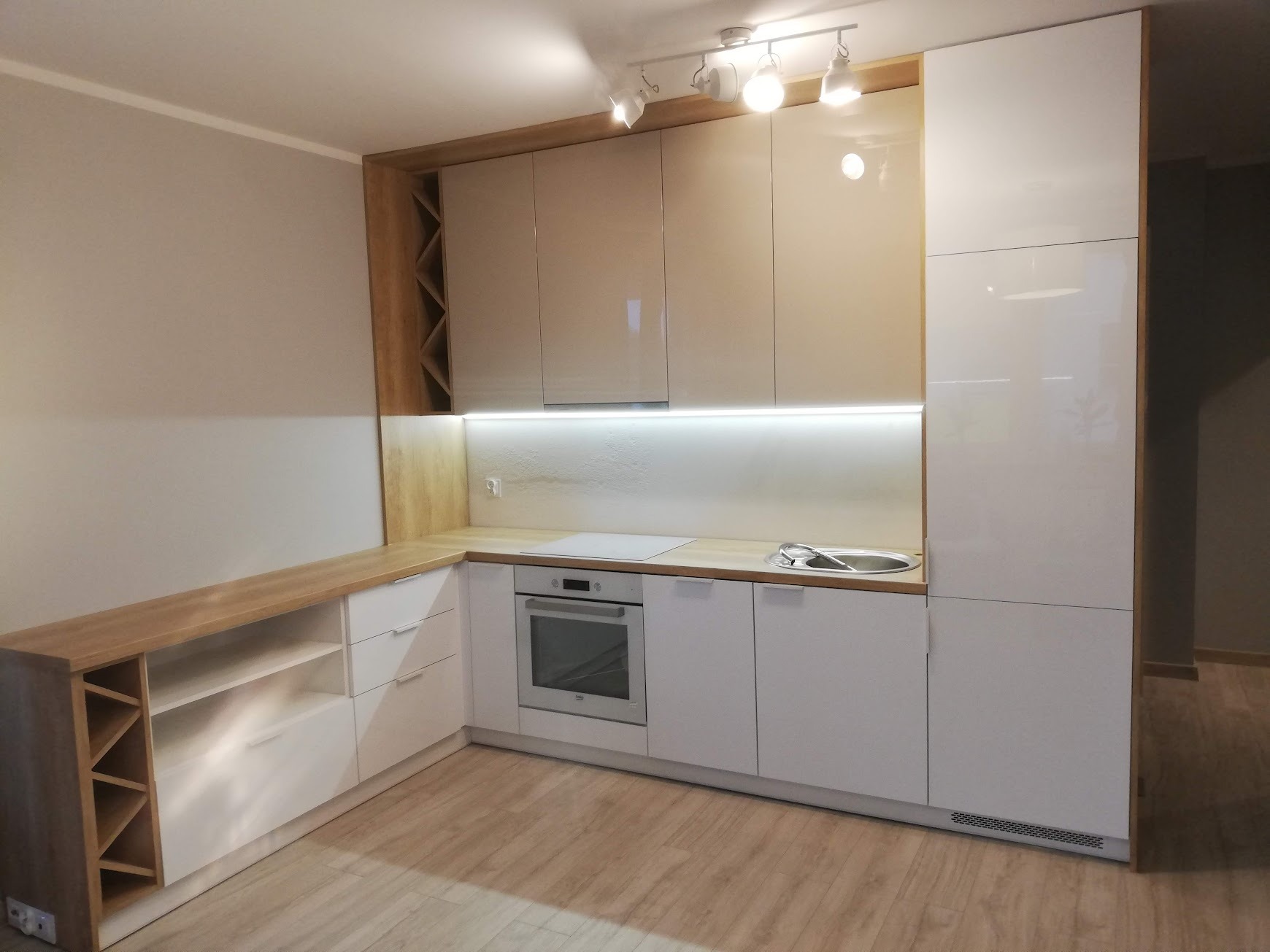 Nowoczesna kuchnia na wymiar z białymi frontami i drewnianym blatem, oświetlenie podszafkowe LED, regał na wino wbudowany w blat.