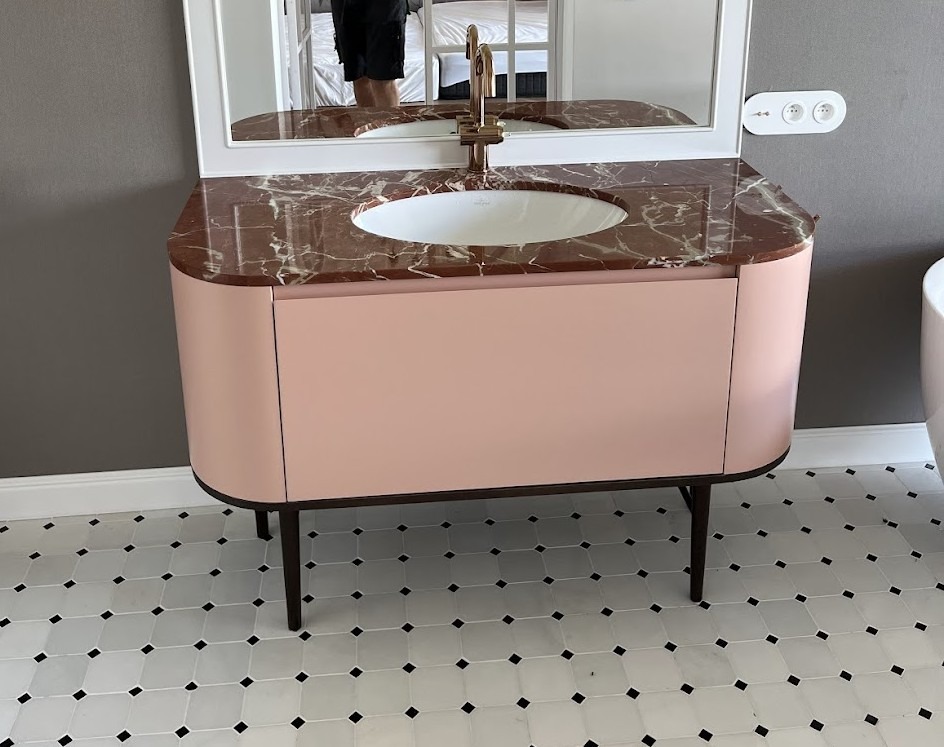 Różowa szafka łazienkowa z marmurowym blatem i złotą armaturą, odbicie osoby w lustrze, biała mozaika podłogowa.