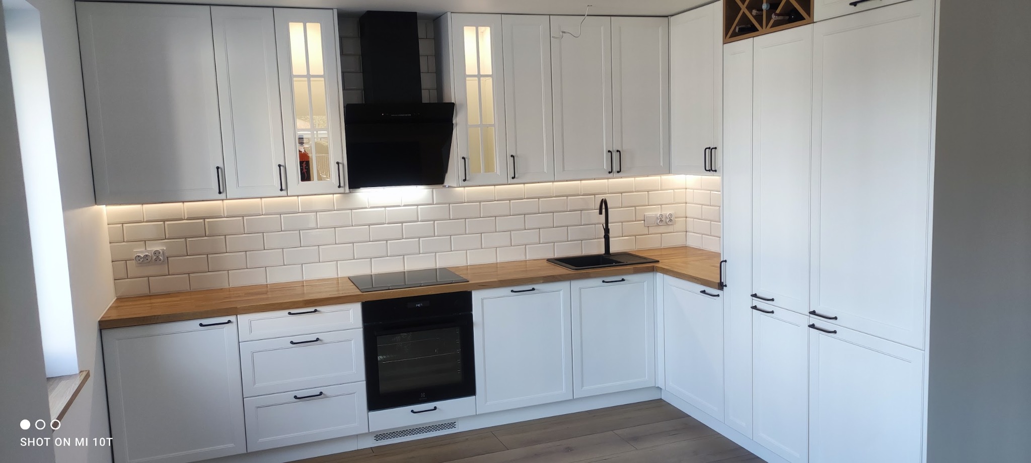 Nowoczesna kuchnia z białymi frontami i drewnianym blatem, czarna armatura, białe płytki metra i podświetlenie LED pod szafkami wiszącymi, widoczny fragment regału na wino.