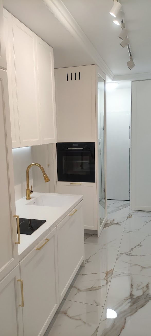 Biała zabudowa kuchenna z piekarnikiem, złotą armaturą i płytą indukcyjną, marmurowe płytki podłogowe.