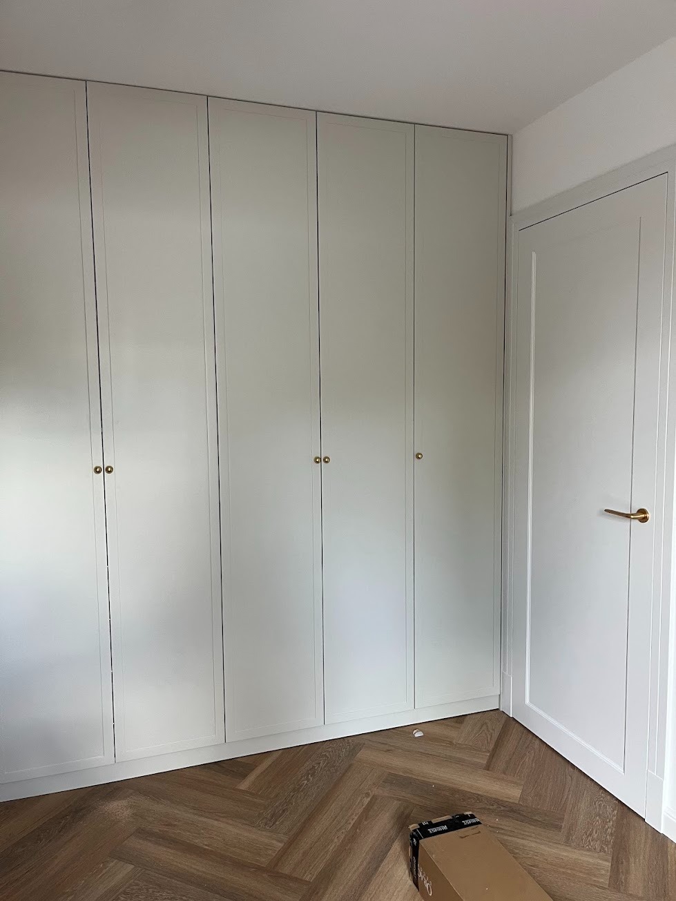 Biała szafa wnękowa z sześcioma drzwiami i złotymi gałkami, dopasowana do koloru drzwi wewnętrznych, na podłodze parkiet w jodełkę, karton po montażu.