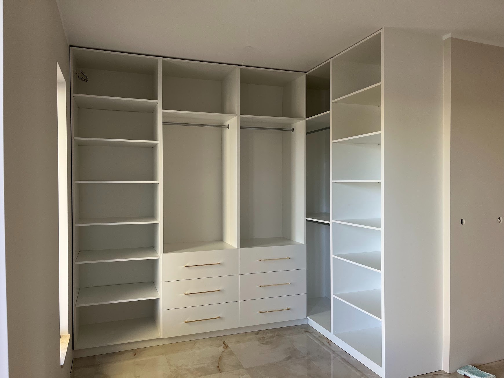 Biała szafa wnękowa z półkami i szufladami z złotymi uchwytami, widoczna w trakcie montażu, z elementami wykończenia wnętrza w tle.