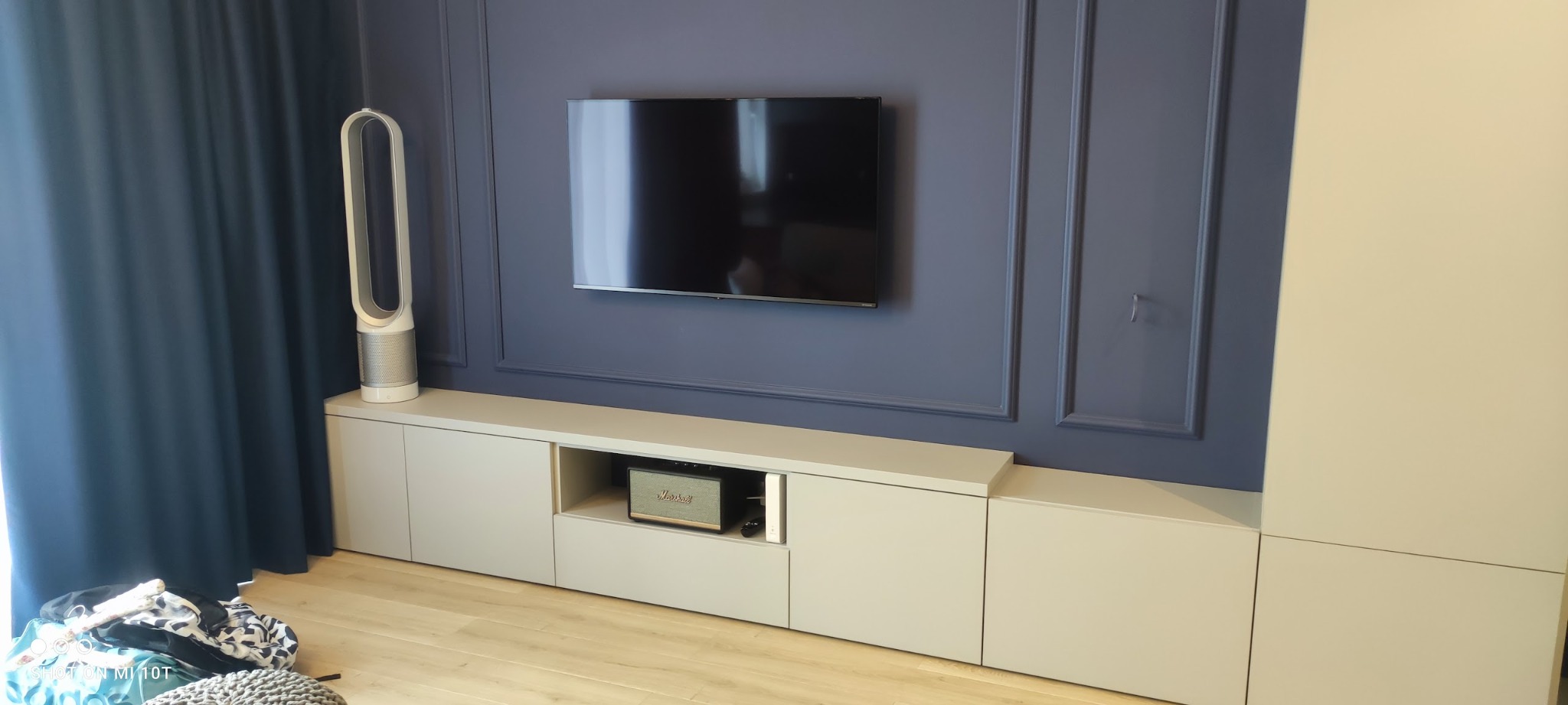 Nowoczesna zabudowa RTV z szarymi frontami, na tle ściany w kolorze granatowym z dekoracyjnymi listwami, widoczny głośnik Marshall i oczyszczacz powietrza Dyson.