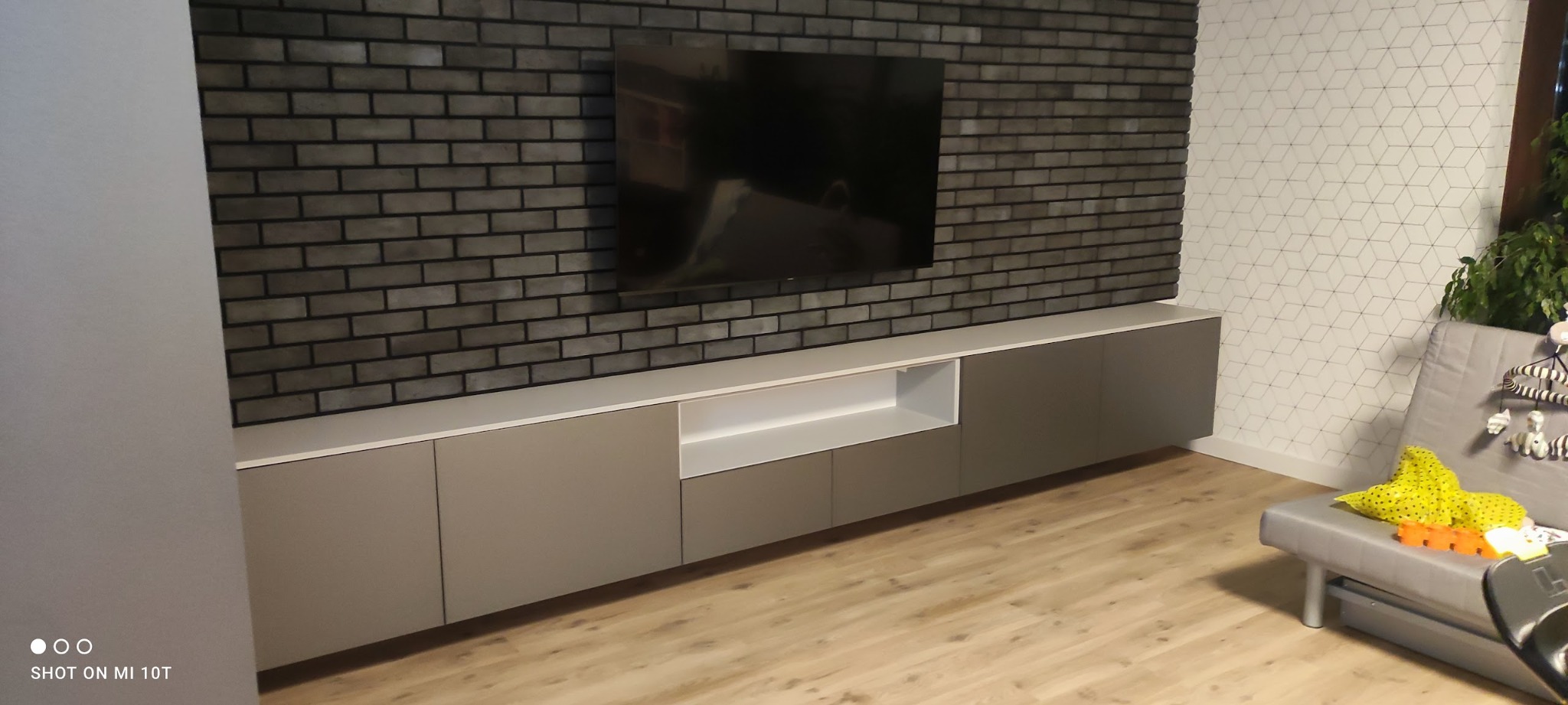 Długa, szara szafka RTV podwieszona pod telewizorem na ścianie z cegły dekoracyjnej, widok z perspektywy podłogi.