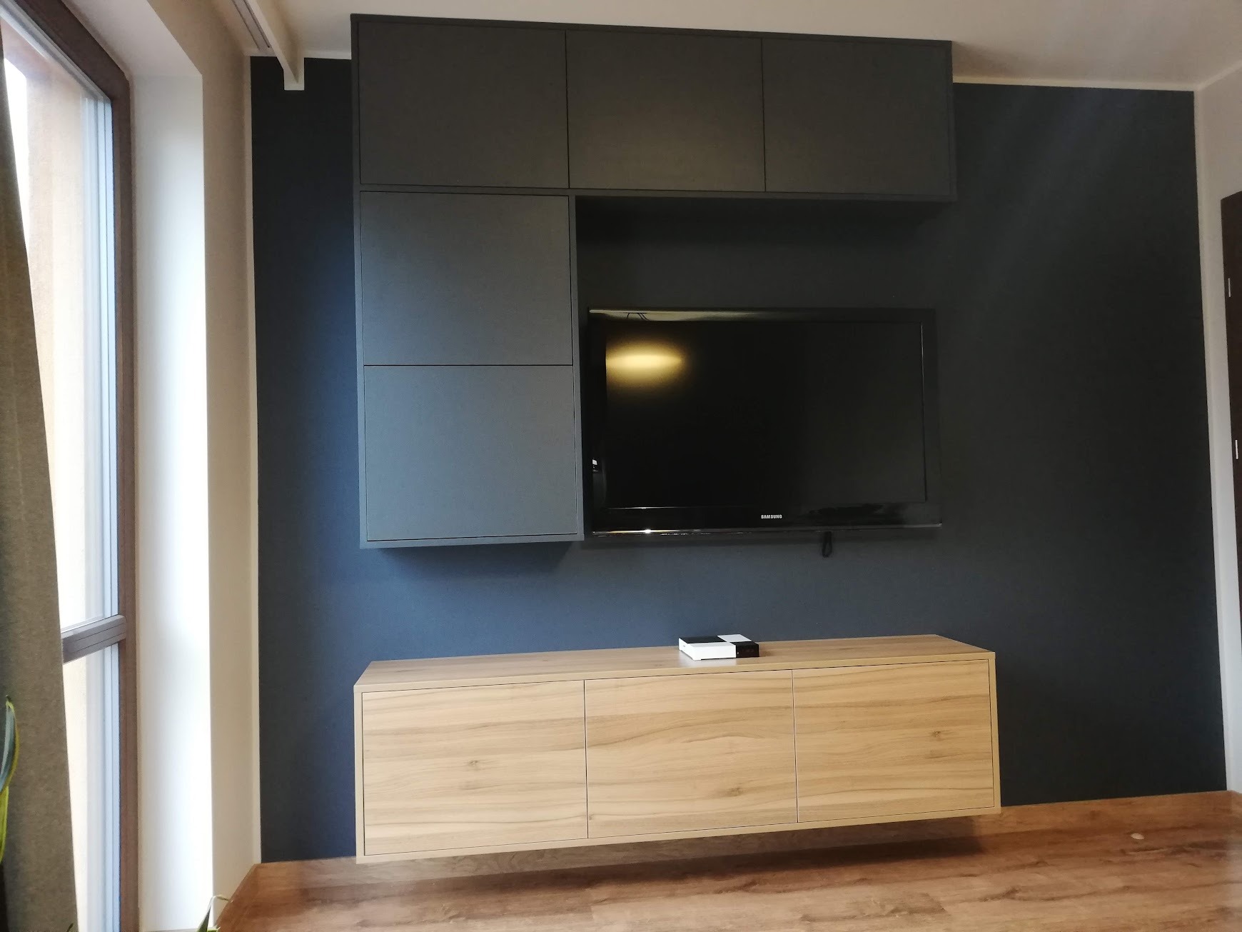 Nowoczesna aranżacja ściany telewizyjnej z szarymi szafkami wiszącymi i podwieszaną szafką RTV z frontami w kolorze jasnego drewna.