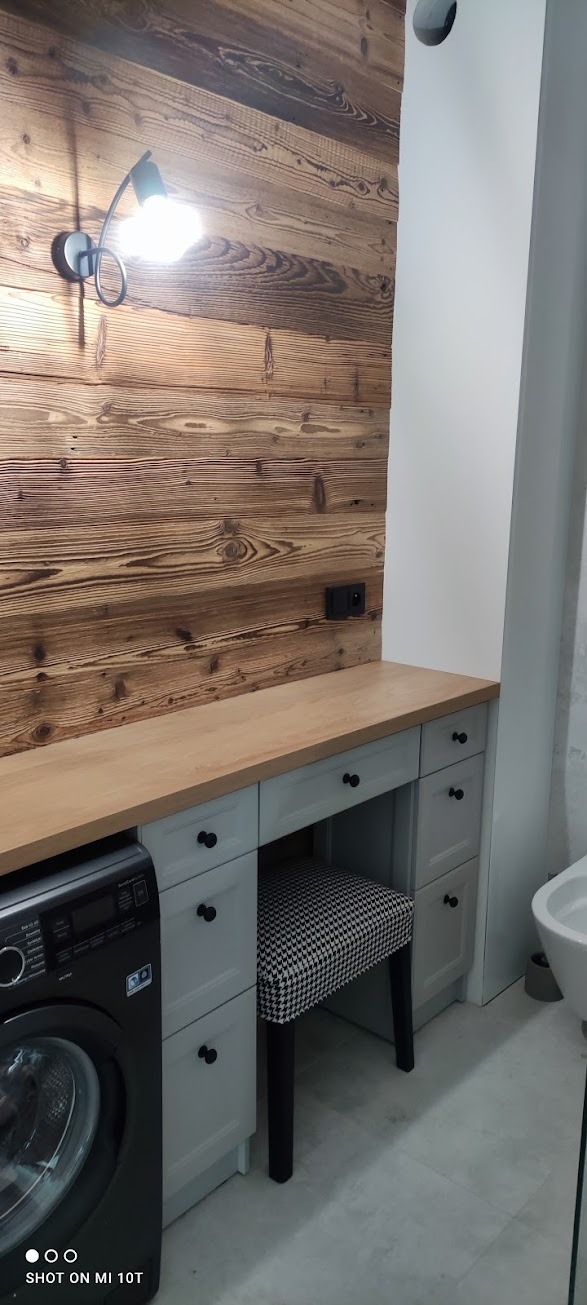 Szara szafka łazienkowa z drewnianym blatem i krzesłem w pepitkę, umieszczona obok pralki na tle ściany z desek i lampy.