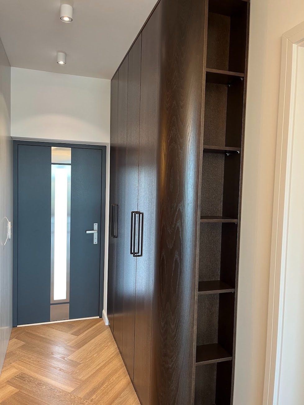 Wysoka, ciemna szafa wnękowa z otwartymi półkami na końcu, z minimalistycznymi, pionowymi uchwytami, w przedpokoju z parkietem w jodełkę i szarymi drzwiami.