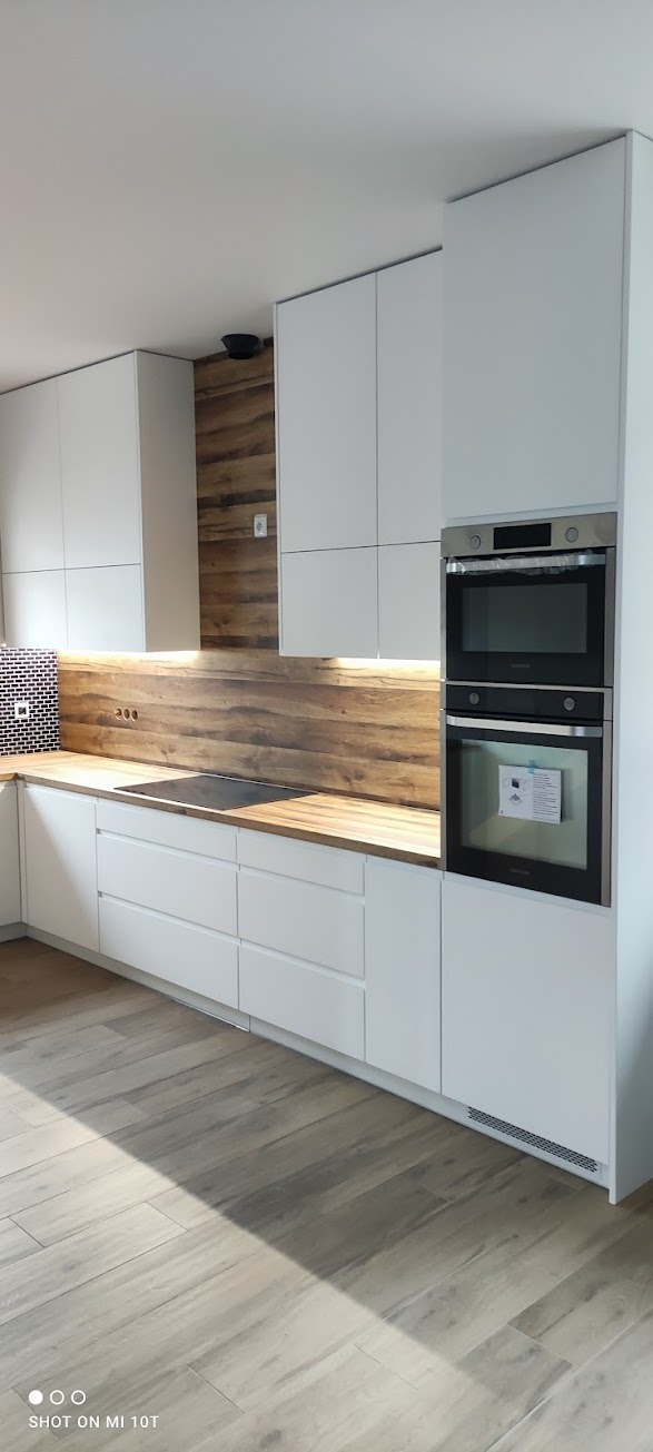 Nowoczesna, minimalistyczna kuchnia z białymi frontami, drewnianym blatem i panelem ściennym imitującym drewno, podświetlonym oświetleniem LED. Widoczny piekarnik w zabudowie i czarna płyta...
