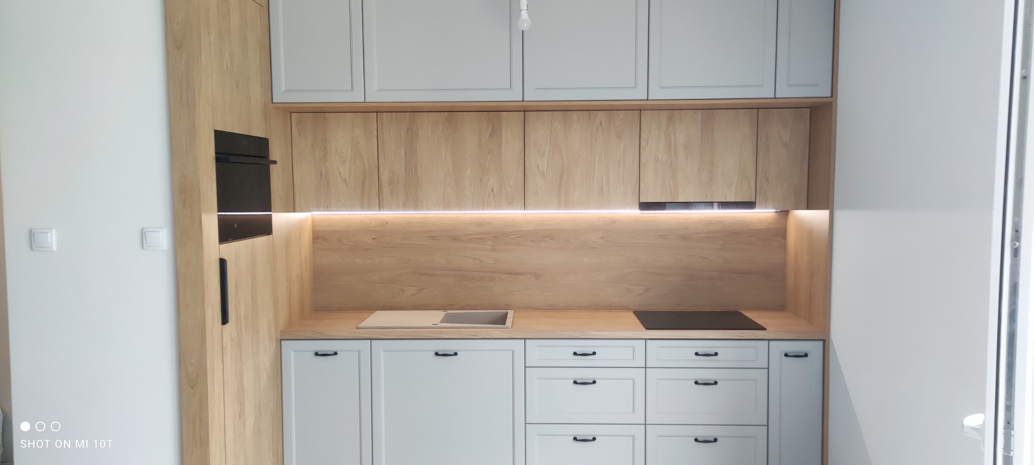 Jasna kuchnia z drewnianymi i szarymi frontami, oświetleniem LED pod szafkami wiszącymi i czarnym piekarnikiem w zabudowie.