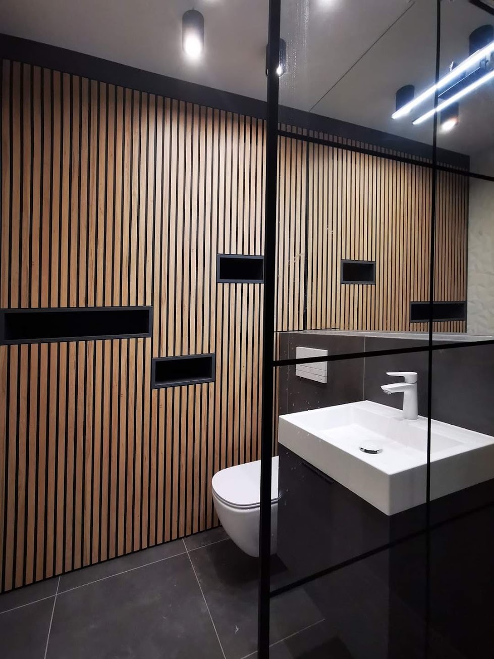 Nowoczesna łazienka z drewnianą ścianą z pionowych lameli z wnękami, białą wiszącą toaletą i umywalką nablatową, oddzielona czarną ramą kabiny prysznicowej.