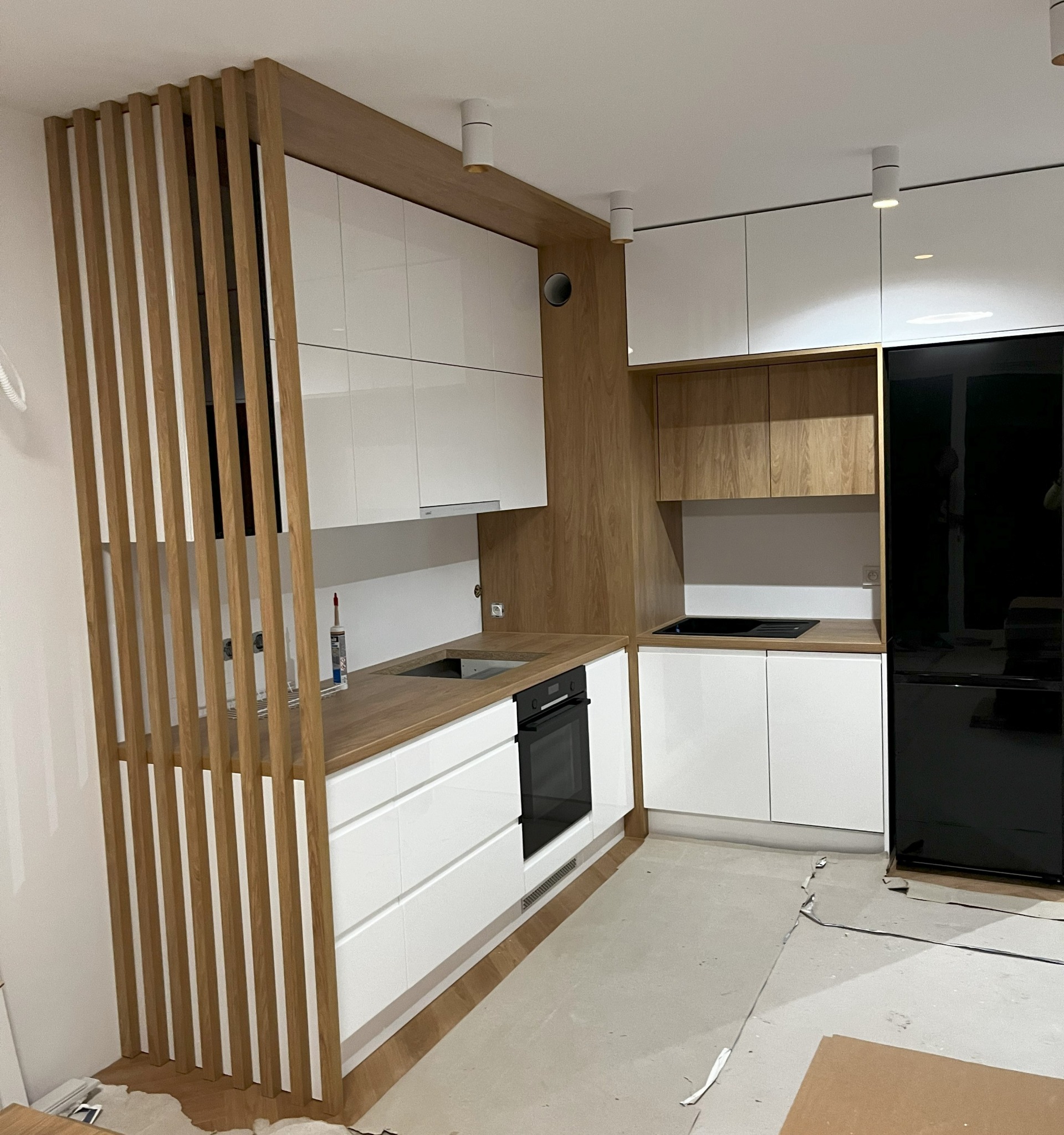 Nowoczesna kuchnia z białymi szafkami i drewnianymi akcentami, widoczna drewniana ścianka działowa z pionowych listew, czarna lodówka i niedokończona podłoga.