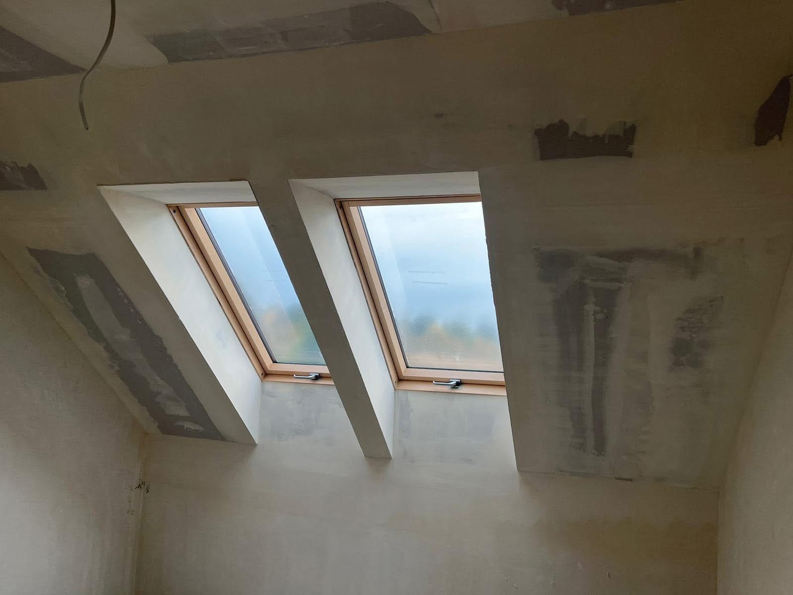 Dwa okna dachowe w surowym stanie, osadzone w skosie poddasza z widocznymi śladami szpachlowania i niedoskonałościami na ścianach i suficie.