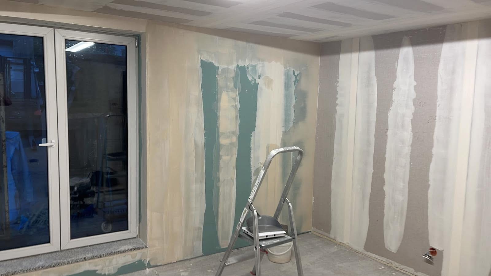 Wnętrze w trakcie montażu płyt karton-gips, widoczne szpachlowane łączenia, drabina aluminiowa i okno z widokiem na zewnątrz.