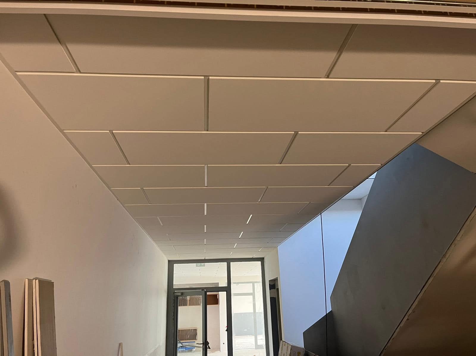 Montaż sufitu podwieszanego z widocznymi płytami i metalową konstrukcją w korytarzu biurowym, widok na drzwi i fragment schodów.