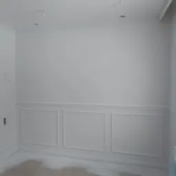 Wnętrze po malowaniu na biało z dekoracyjnymi panelami ściennymi i listwami przysufitowymi, widoczne kable wystające z sufitu, podłoga częściowo zakryta folią malarską.