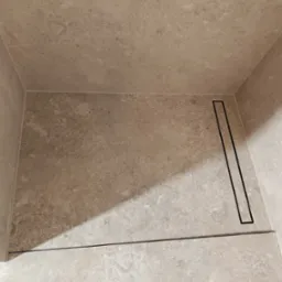 Wykończony brodzik z beżowych płytek imitujących kamień z liniowym odpływem w rogu kabiny.