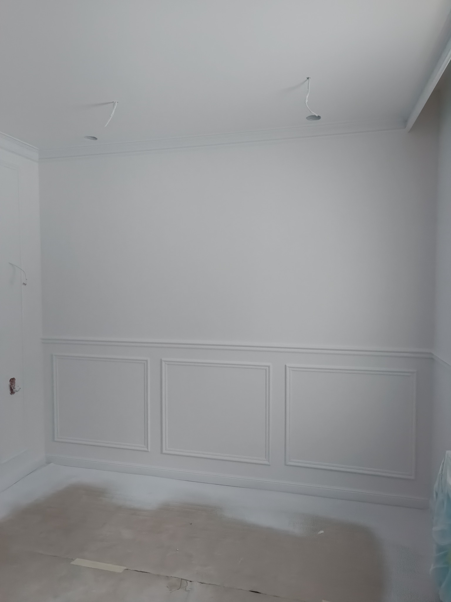 Wnętrze po malowaniu na biało z dekoracyjnymi panelami ściennymi i listwami przysufitowymi, widoczne kable wystające z sufitu, podłoga częściowo zakryta folią malarską.