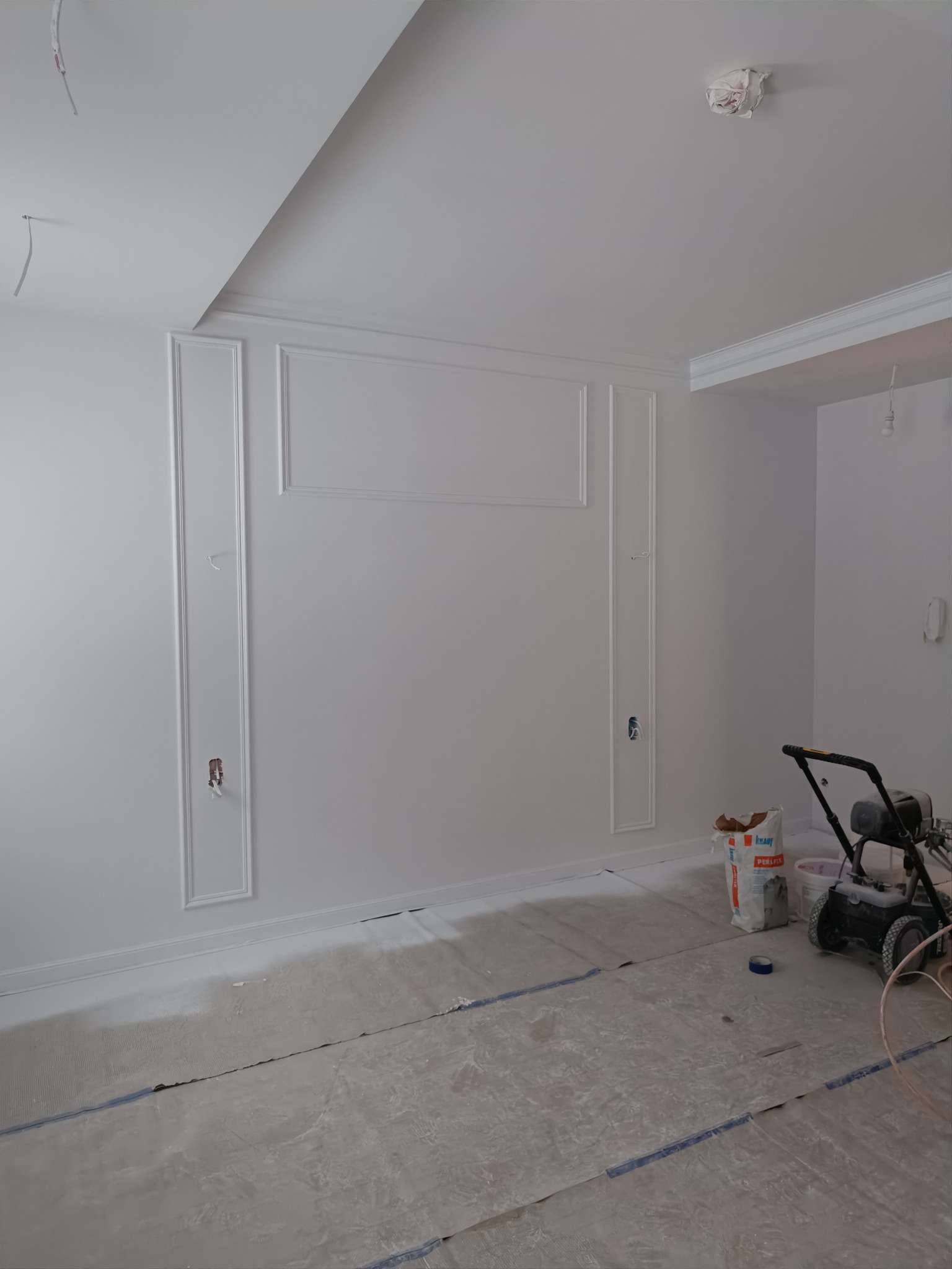 Białe ściany z dekoracyjnymi listwami i gładzią, widoczny agregat malarski i worek gładzi na zabezpieczonej podłodze, gniazdka elektryczne częściowo odsłonięte.