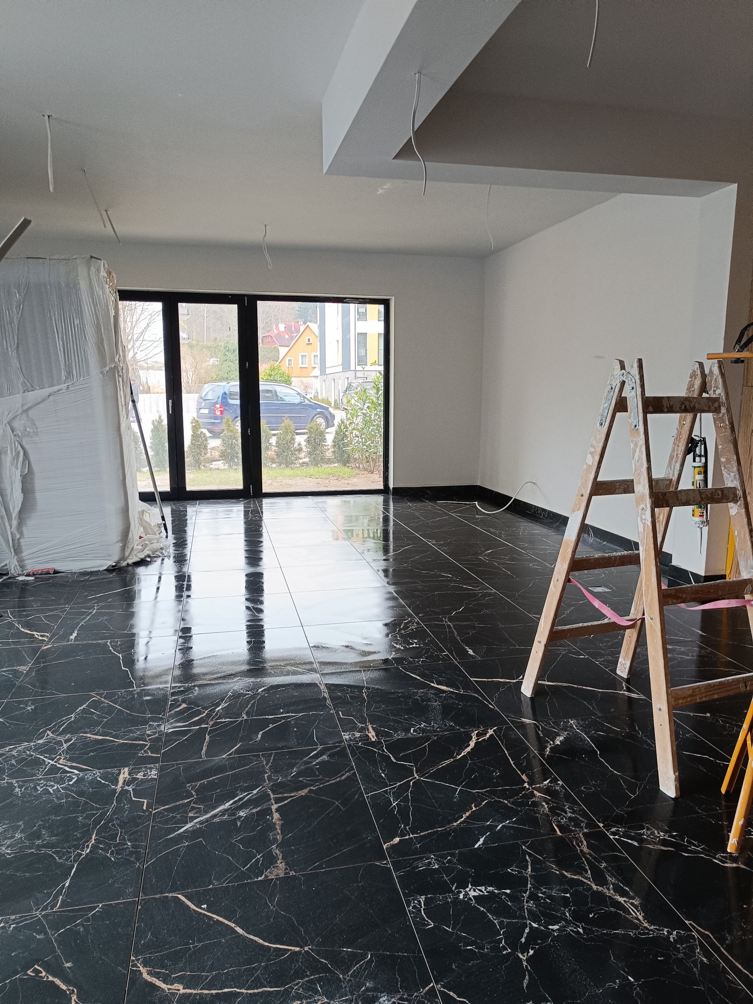 Błyszcząca, czarna podłoga z marmurkowym wzorem w nowym wnętrzu, drabina oparta o ścianę, okno z widokiem na domy i samochód, zakryty folią element wyposażenia.