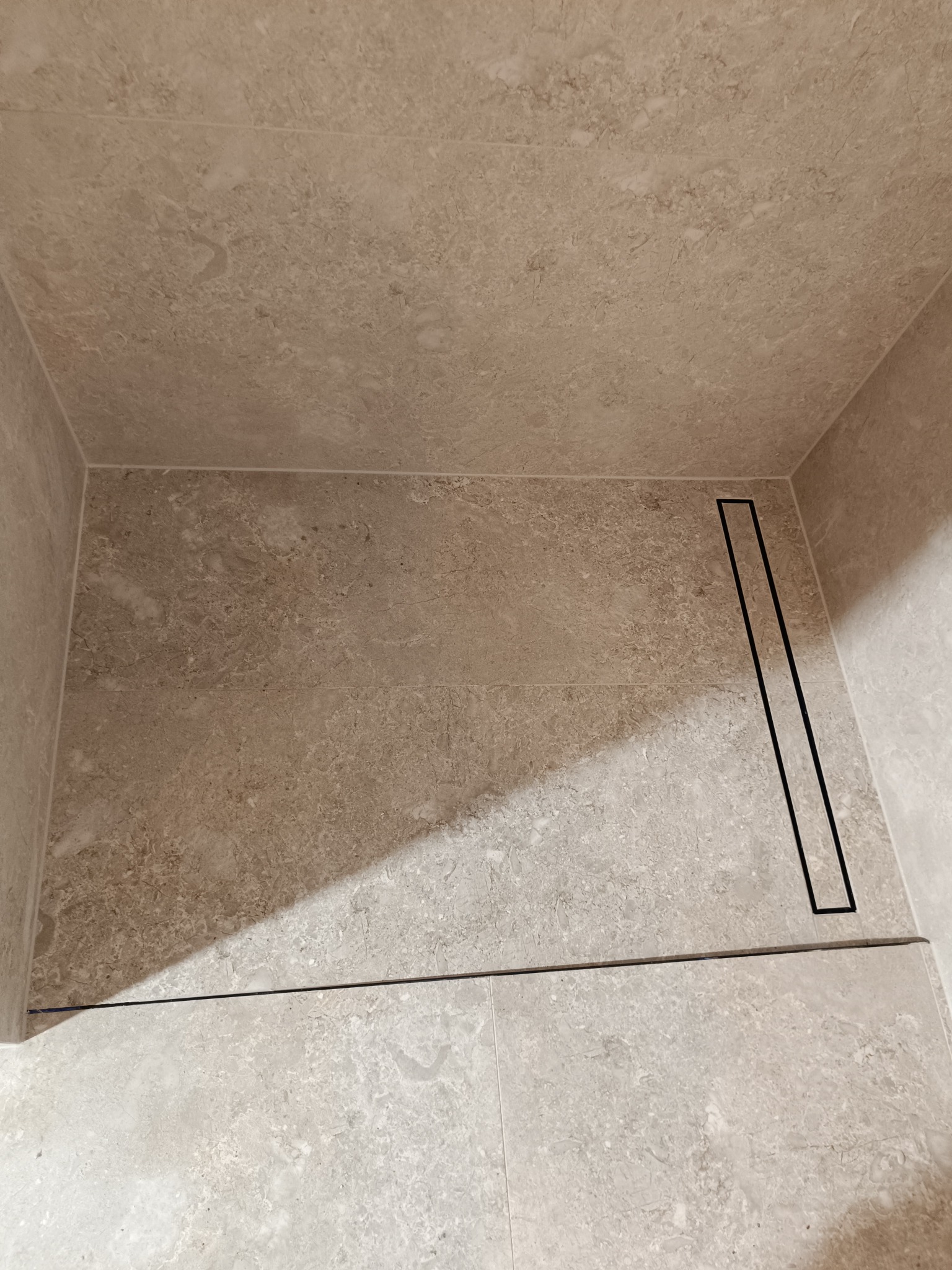 Wykończony brodzik z beżowych płytek imitujących kamień z liniowym odpływem w rogu kabiny.