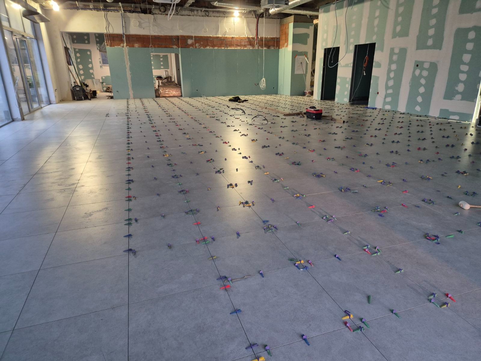 Szare płytki podłogowe w trakcie układania, widoczne liczne plastikowe krzyżyki dystansowe w różnych kolorach, ściany w surowym stanie z płytami gipsowo-kartonowymi, fragmenty instalacji...