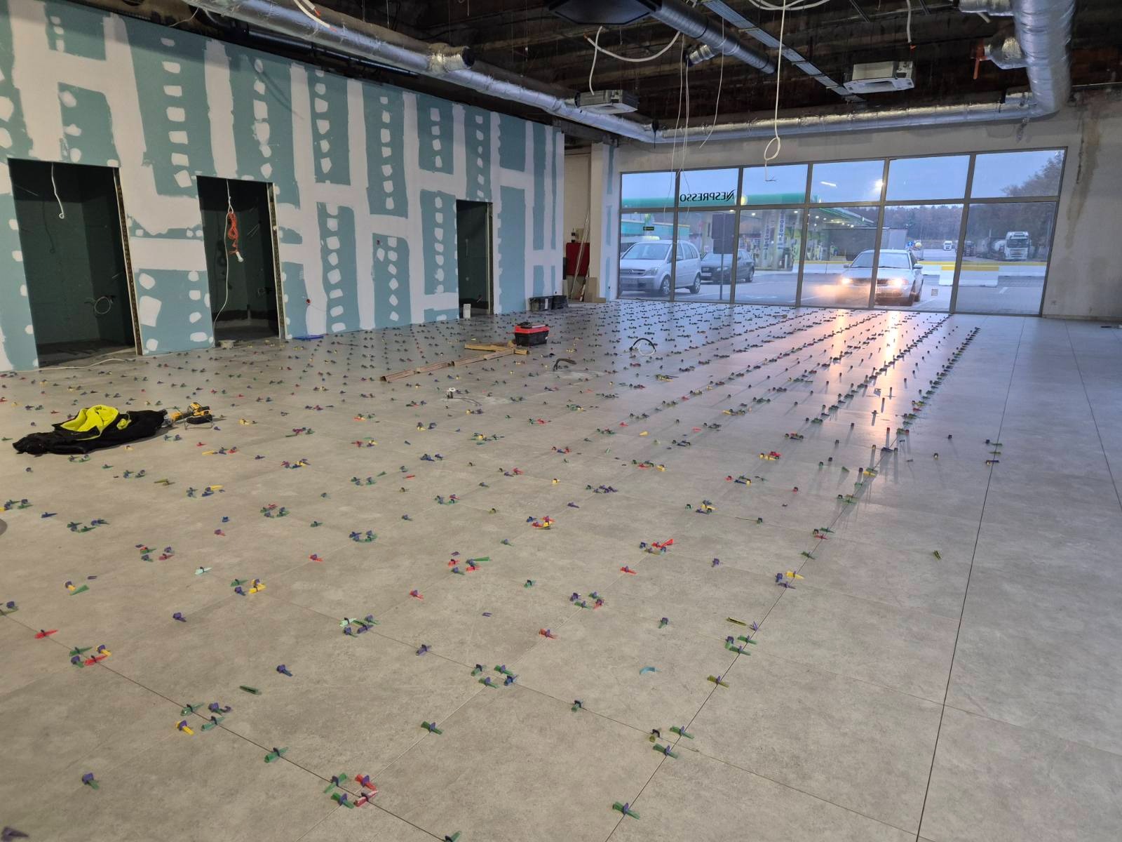 Szare płytki podłogowe w trakcie układania, z systemem poziomowania w postaci kolorowych klinów i klipsów, widoczne przygotowane ściany z otworami drzwiowymi oraz okno z widokiem na stację benzynową