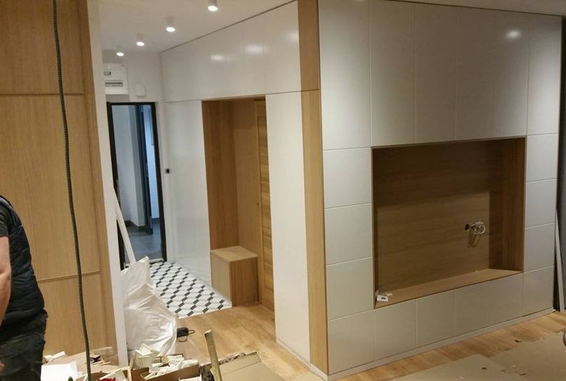 Wnęka w zabudowie z białymi frontami i drewnianym wnętrzem, widoczne kable instalacji elektrycznej, podłoga w trakcie montażu, widoczne narzędzia i materiały budowlane, fragment sylwetki osoby...