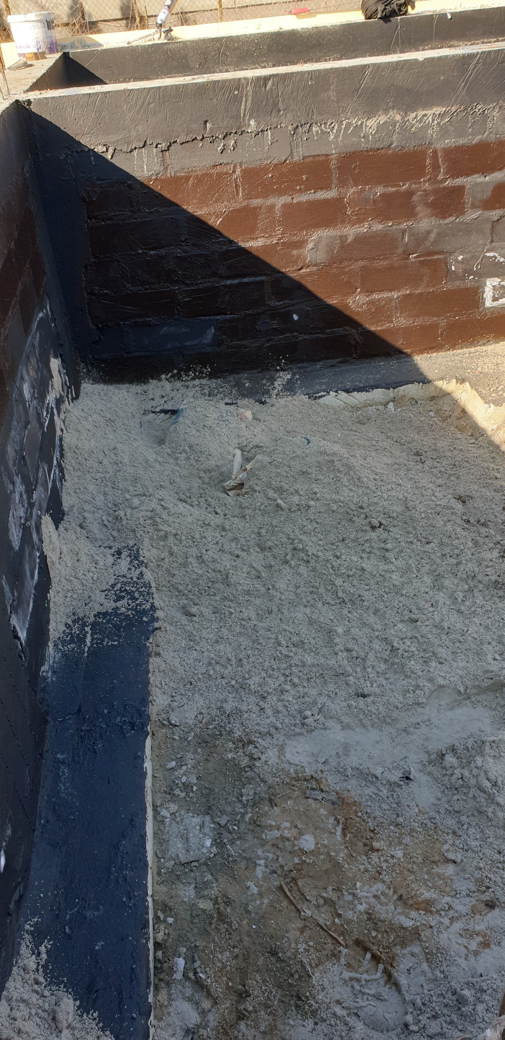 Murowana ściana z cegły pomalowana na brązowo, z czarną hydroizolacją na górze oraz warstwą piasku lub zaprawy na dole, rzucająca cień na część muru.