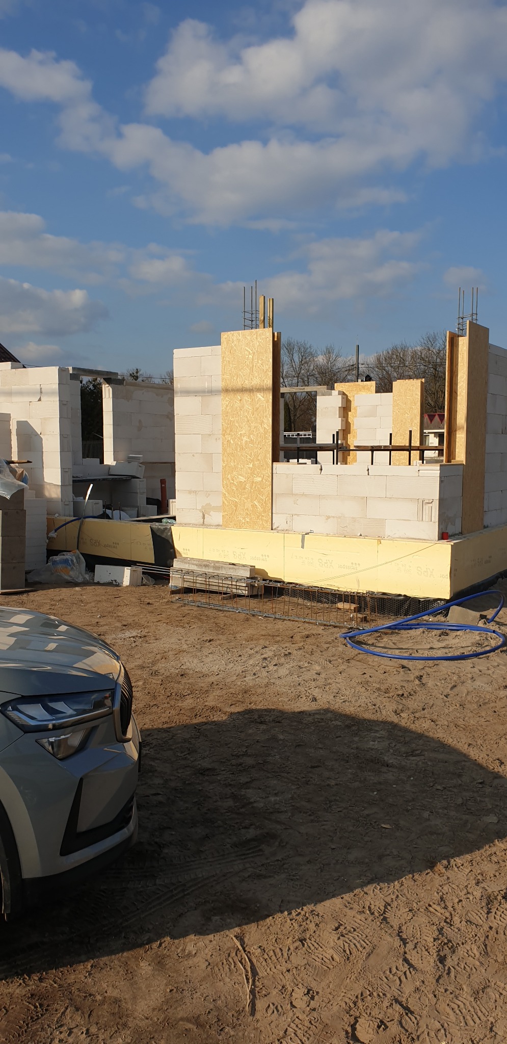 Budowa domu w trakcie wznoszenia ścian z bloczków, widoczne szalunki z płyty OSB i zbrojenie fundamentów, obok stoi samochód osobowy.