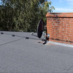 Antena satelitarna zamontowana na dachu obok ceglanego komina, z zielonymi drzewami w tle i błękitnym niebem. Widok z góry na szary dach pokryty papą.