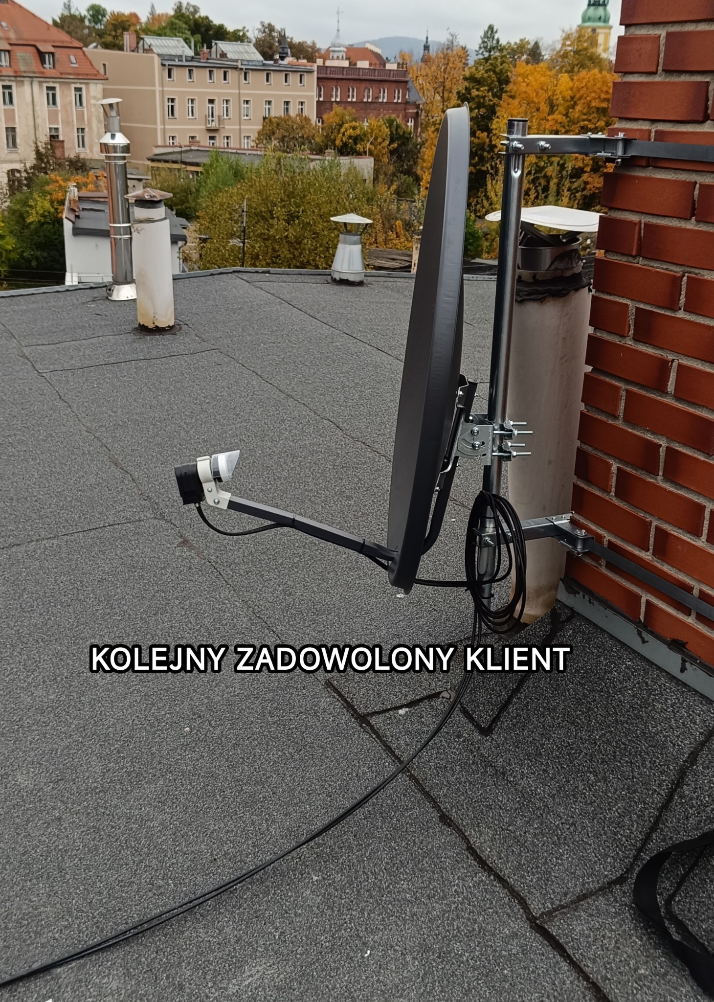 Antena satelitarna na dachu budynku z ceglaną ścianą, widok na zabudowę miejską w tle. Kabel antenowy częściowo widoczny na dachu. Zamontowana antena.