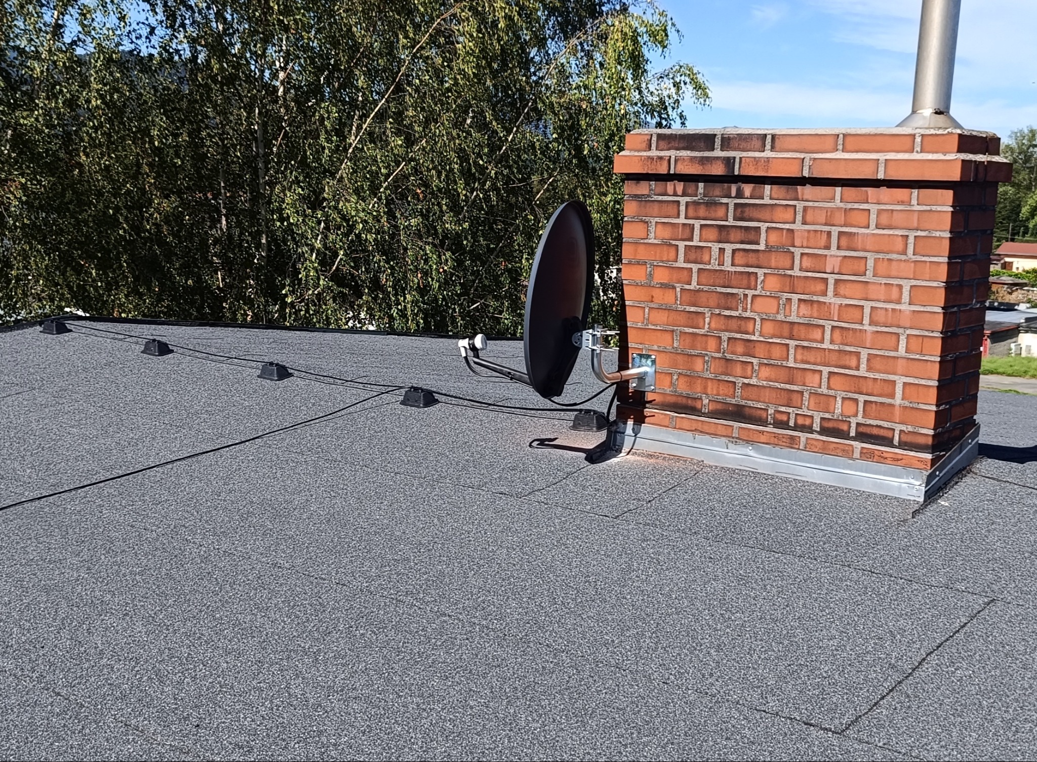 Antena satelitarna zamontowana na dachu obok ceglanego komina, z zielonymi drzewami w tle i błękitnym niebem. Widok z góry na szary dach pokryty papą.