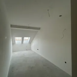 Wnętrze poddasza po gładziowaniu ścian i sufitu, widoczne dwa okna dachowe i wystające przewody elektryczne. Szare wykończenie podłogi.